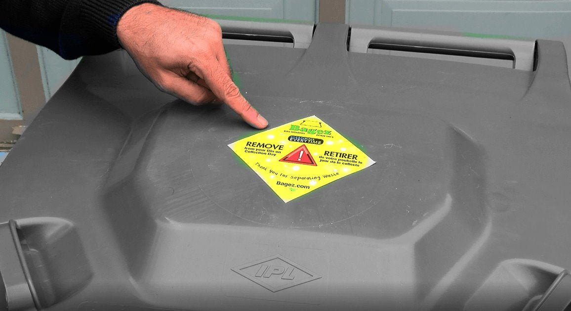 Hand pointing to a bin lid sticker, a yellow warning label on a gray trash can lid